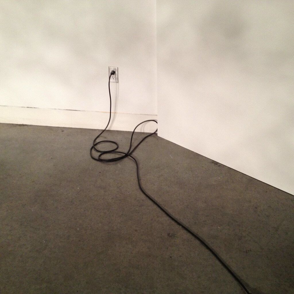 An ordinary corner. Before it’s “a room,” it’s a dark floor, a pale wall and a loop of black cable.